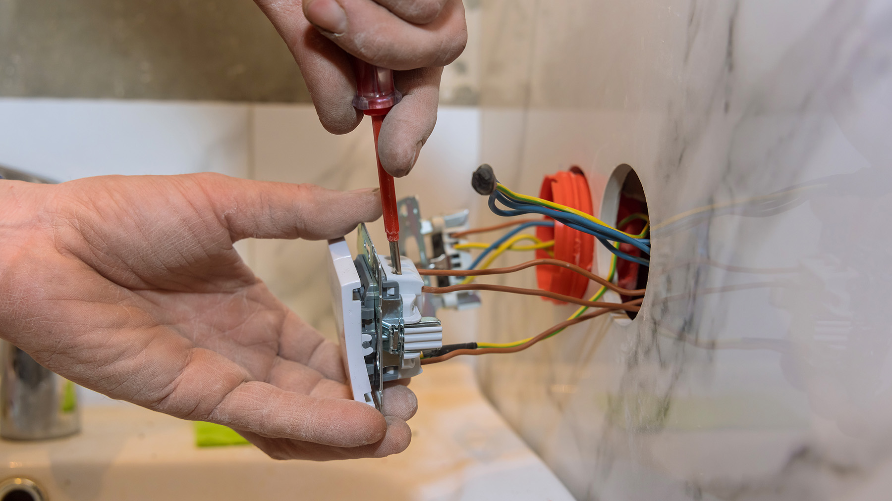 A hand holding a small electronic device with visible circuitry and connectors, being worked on by someone in front of a marble wall.