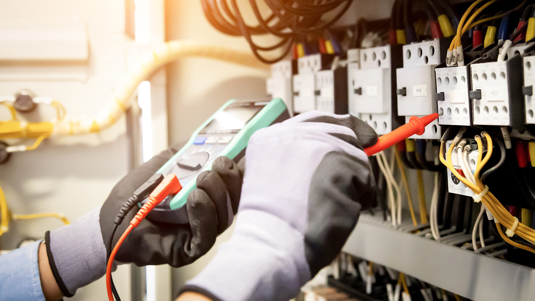 The image depicts a person wearing gloves handling electrical wiring and connectors at a panel with various switches and cables, set against an industrial background.