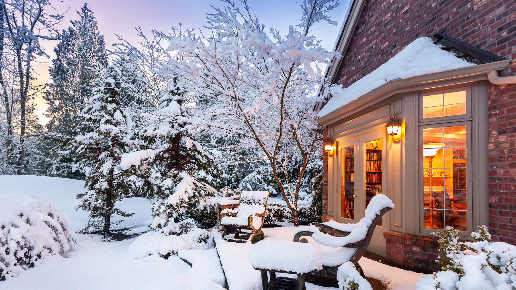 The image depicts a snowy residential scene featuring a house with a lit porch, surrounded by trees with snow on their branches.
