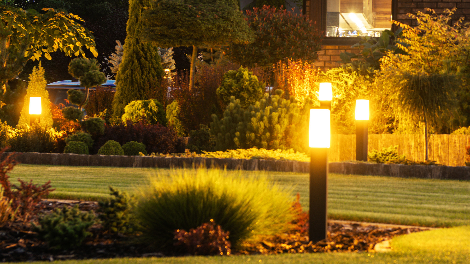 The image shows a well-maintained garden at night with illuminated outdoor lights, pathway lamps, and a neatly trimmed lawn.
