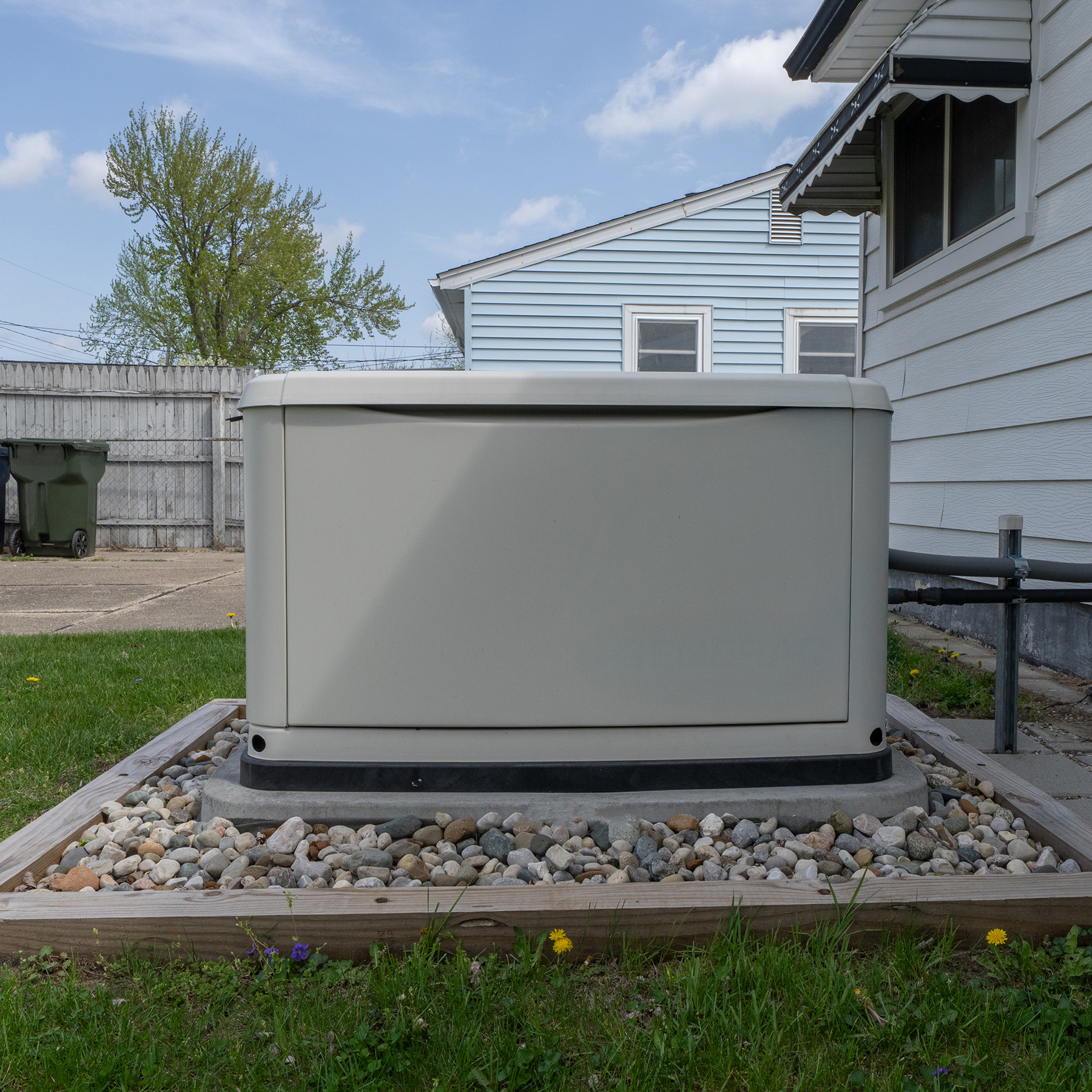 The image shows a large, gray outdoor utility box with a black lid, placed on a gravel bed surrounded by small rocks, situated in front of a house with a white exterior and a garage door.