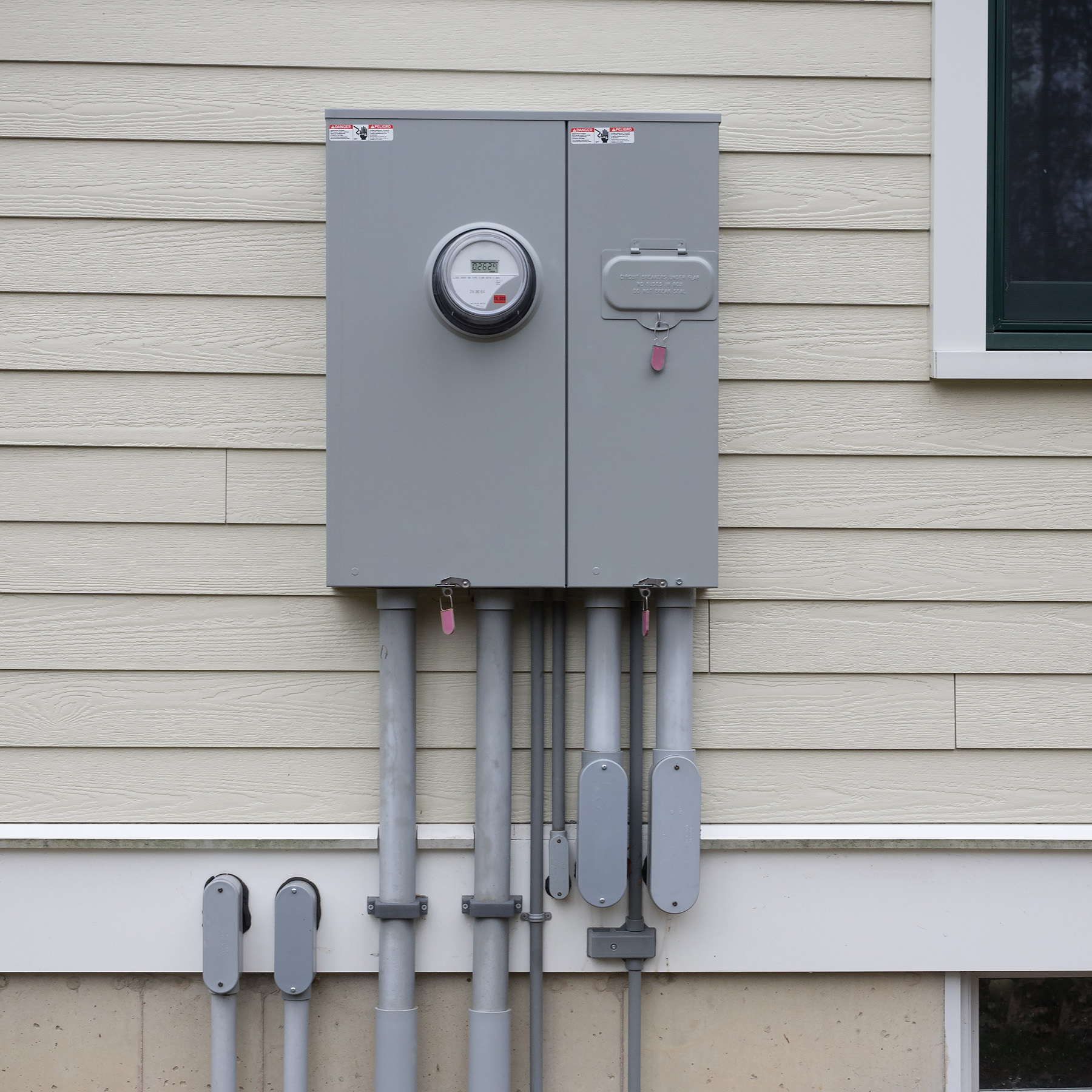 The image features an electric meter attached to a gray utility box with a digital display on a wall adjacent to a beige house, indicating a residential setting with electrical infrastructure.