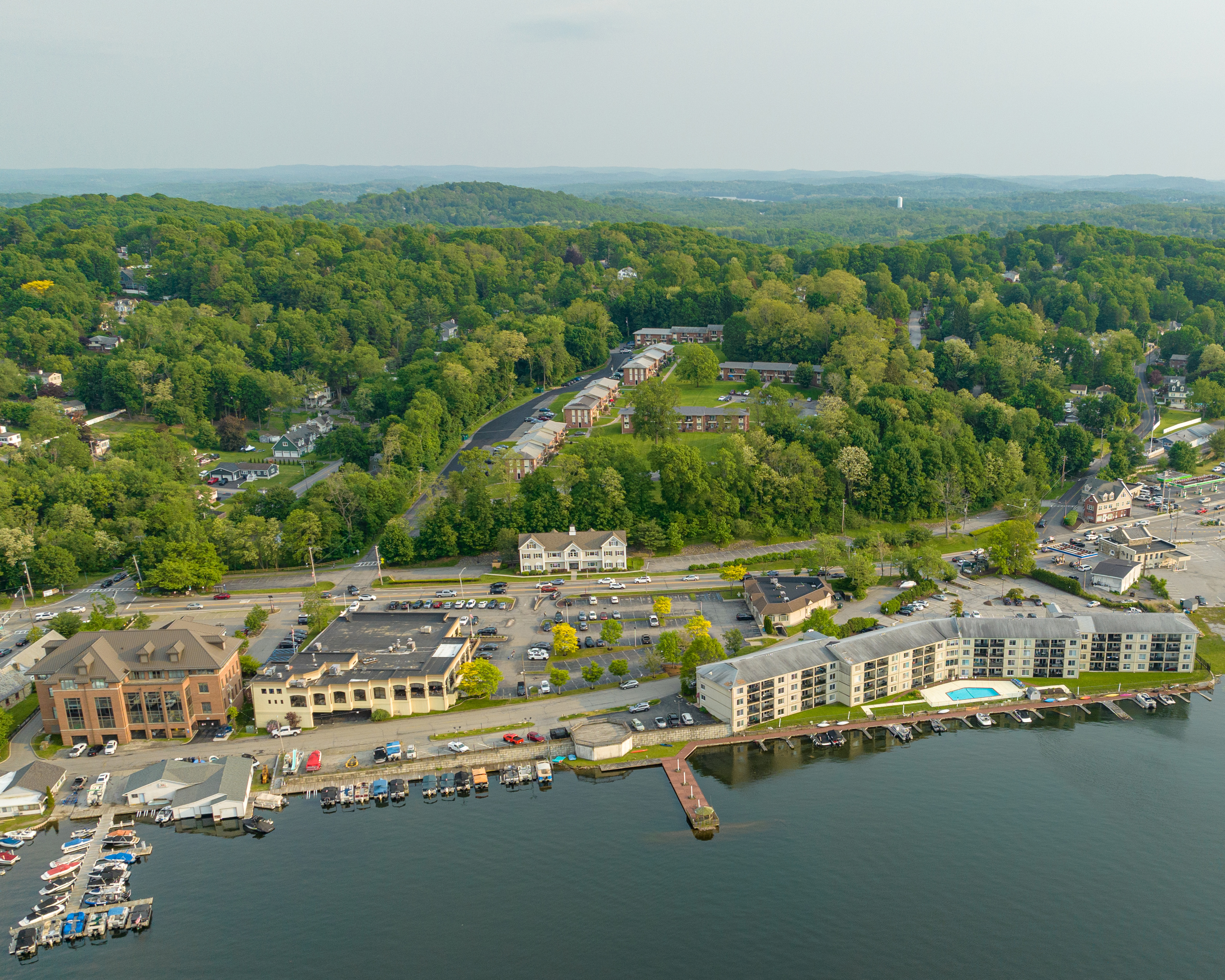 This is a color photograph depicting an aerial view of a suburban area with a mix of residential houses, roads, and green spaces, surrounded by water and hills, on a clear day.