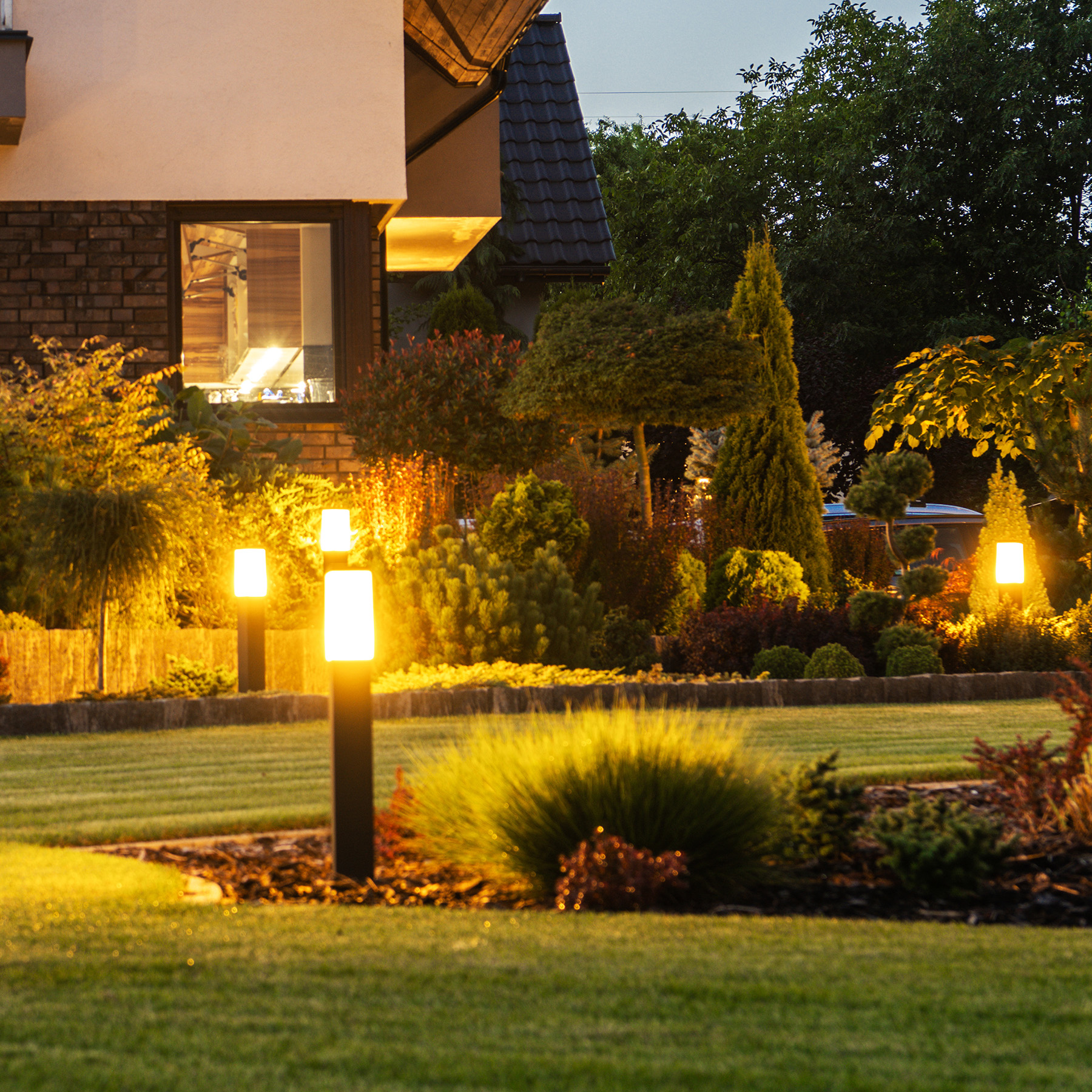 The image shows a well-maintained residential garden at night with illuminated path lights, shrubs, and a house with exterior lighting.