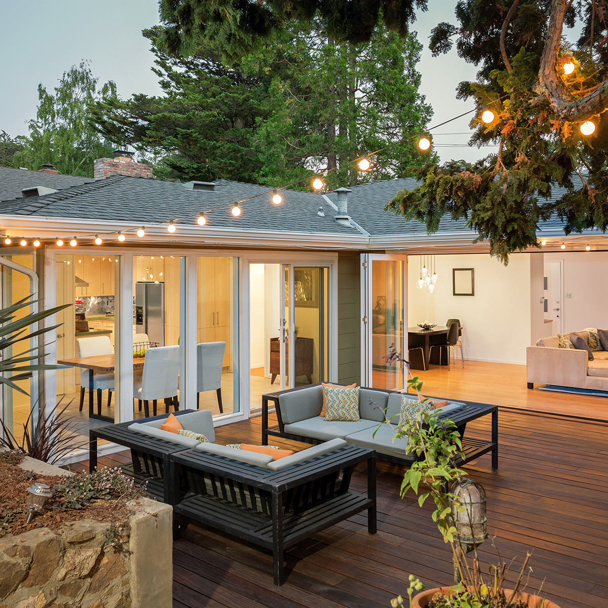 The image depicts a modern outdoor living space with a covered patio, wooden decking, a pergola-covered seating area with a sofa and coffee table, a dining table surrounded by chairs, a string of lights overhead, a backyard lawn, and a two-story house in the background.