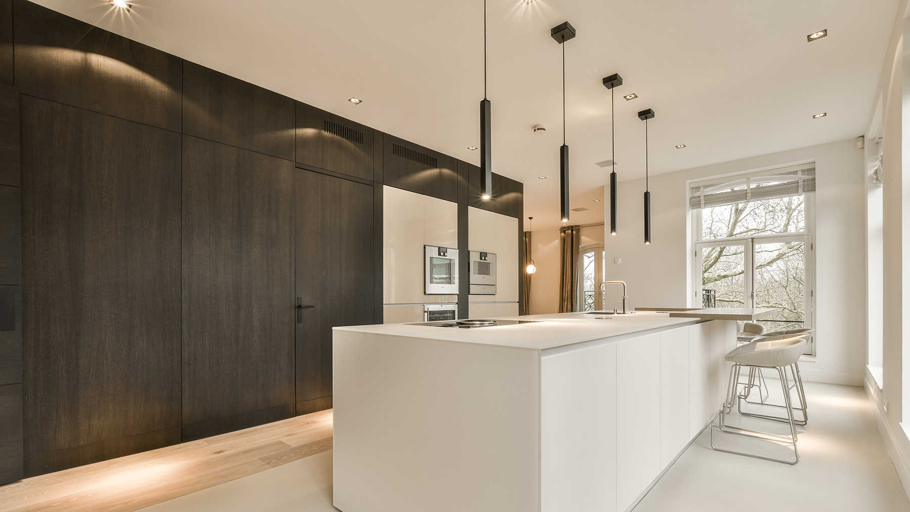 The image depicts a modern kitchen interior with dark wood paneling, white countertops, stainless steel appliances, hanging pendant lights, and a large island with a built-in stove.